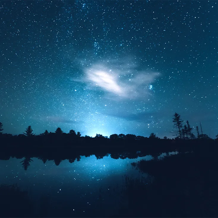Ciel de nuit rempli d'étoiles qui se reflètent sur un lac calme entouré d'une forêt sombre.