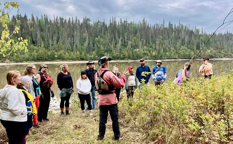 Groupe de personnes au bord d'un lac écoutant un guide en tenue traditionnelle dans une forêt de sapins.