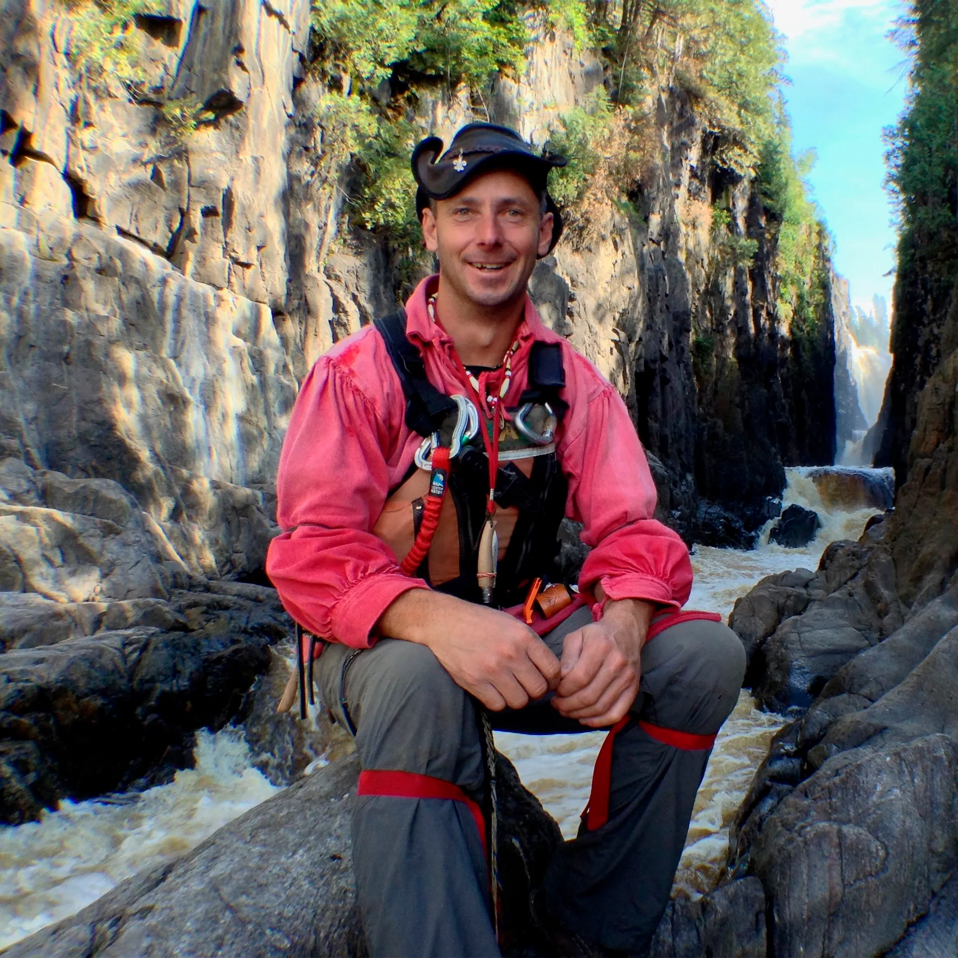 Photo d’André Bernier, souriant, assis sur un rocher. Derrière lui, on voit une rivière sauvage qui coule entre de grandes falaises.