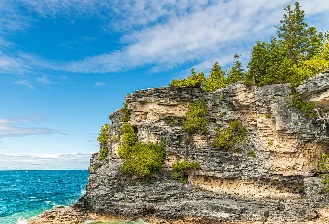 Une haute falaise de pierre grise et beige au bord d’une eau bleue, avec des sapins verts au sommet.