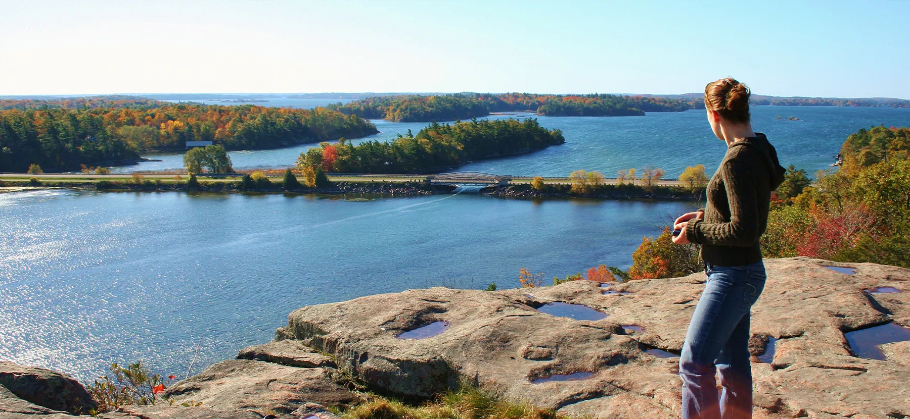 Une femme regarde de grands lacs et des petites îles boisées depuis une haute roche plate.