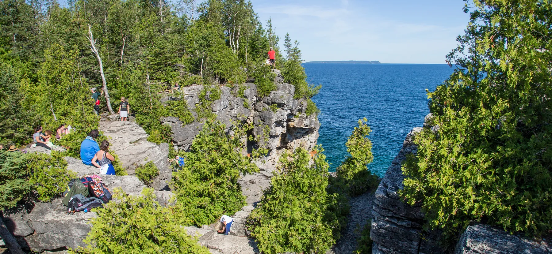 Des randonneurs marchent sur une haute falaise rocheuse entourée d’arbres verts au-dessus d’un grand lac bleu.