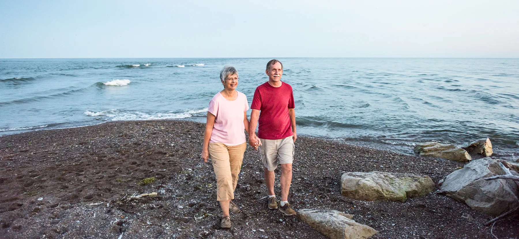Un homme et une femme âgés sourient en marchant main dans la main sur une plage de petits cailloux au bord d'un grand lac.