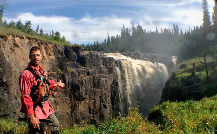 André Bernier sourit devant une grande cascade sauvage entourée de falaises et de sapins.