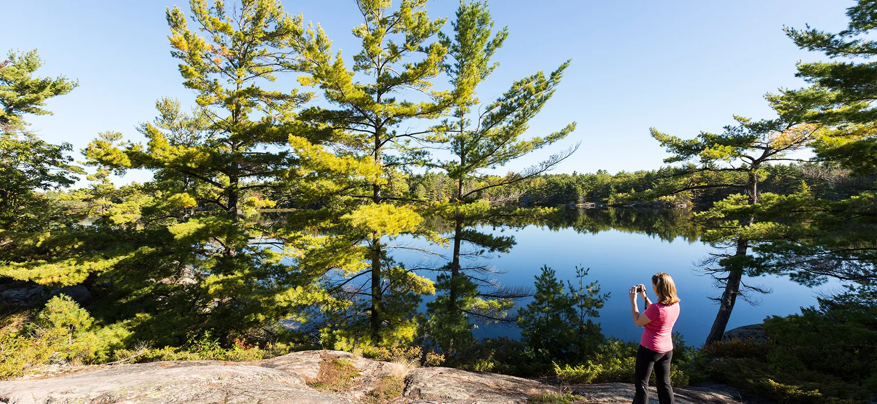 Une femme de dos prend en photo un lac calme entouré de grands sapins verts depuis une roche.